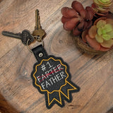 Close‑up of embroidered #1 Father keychain with crossed‑out text
