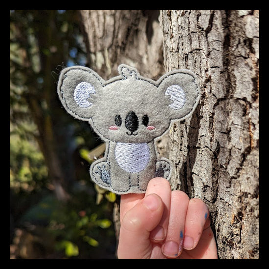 Child's hand with a handmade grey koala finger puppet on their index finger resting in front of a tree.
