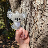 Child's hand with a handmade grey koala finger puppet on their index finger resting in front of a tree.
