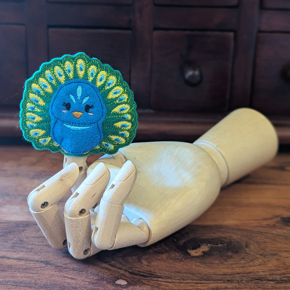Embroidered peacock finger puppet made from felt displayed on a wooden mannequin hand. 