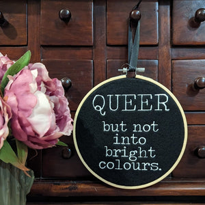 Black and white machine embroidery mounted inside a bamboo hoop with typewriter style text which reads " QUEER but not into bright colours"
