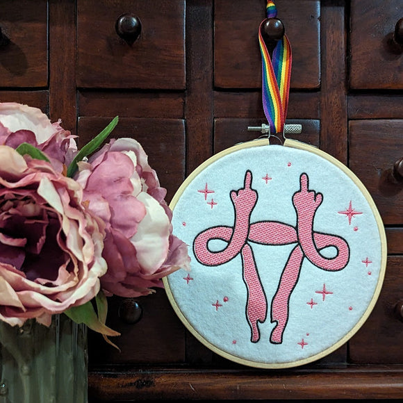 Machine emboidery mounted onto a bamboo hoop depicting a uterus holding up it's fallopian tubes like arms with middle fingers raised. 