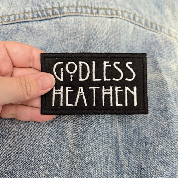 A hand hovering over a light denim jacket holding a black felt iron on patch with the text Godless Heathen embroidered in large white letters.