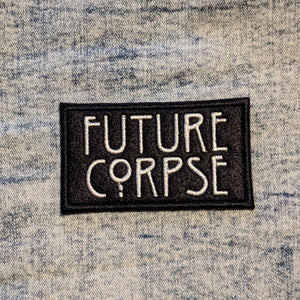 A black felt iron on patch with the text Future Corpse embroidered in large white letters sitting on light denim fabric.