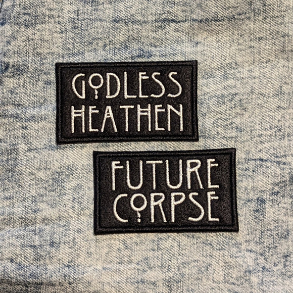 A pair of black felt iron on patches, one above the other with large white text that read Godless Heathen and Future Corpse. Patches sit on a light denim fabric background.