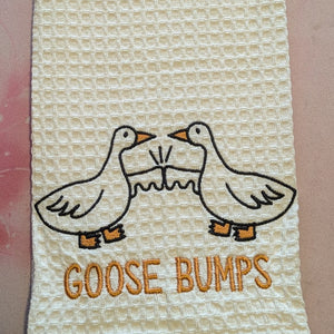 A white cotton waffle weave tea towel embroidered with two geese holding out their wings in a fist bump gesture with the words "goose bumps" underneath.