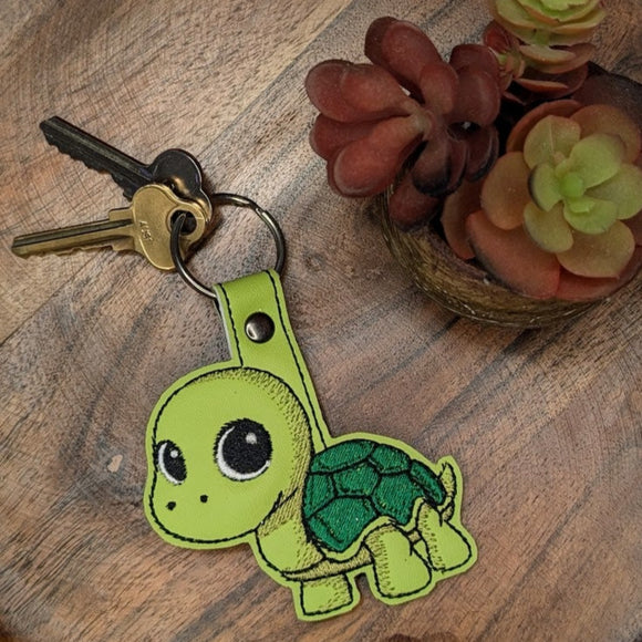 Green turtle keychain on a wooden tray with small plants