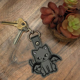 Close‑up of embroidered grey winged cat keychain on faux leather